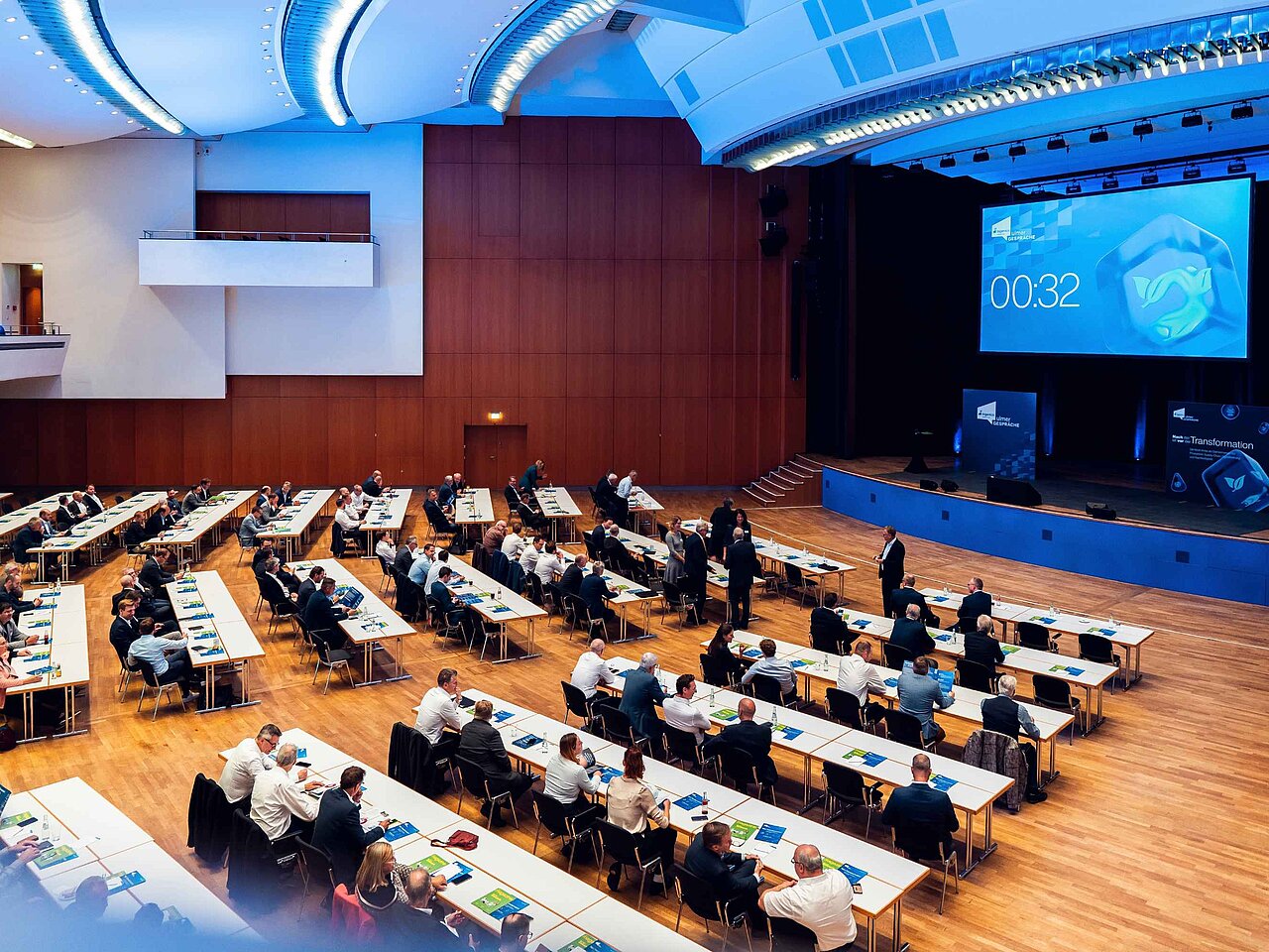 Teilnehmende einer Fachveranstaltung sitzen im Konferenzsaal vor einer Bühne mit Präsentationsbildschirm