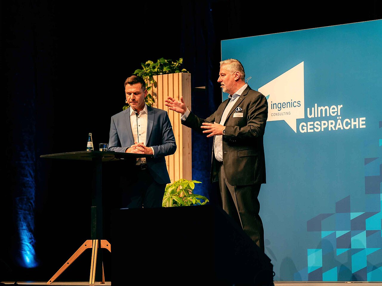 Zwei Referierende diskutieren auf der Bühne der Ulmer Gespräche vor einer Veranstaltungswand von Ingenics
