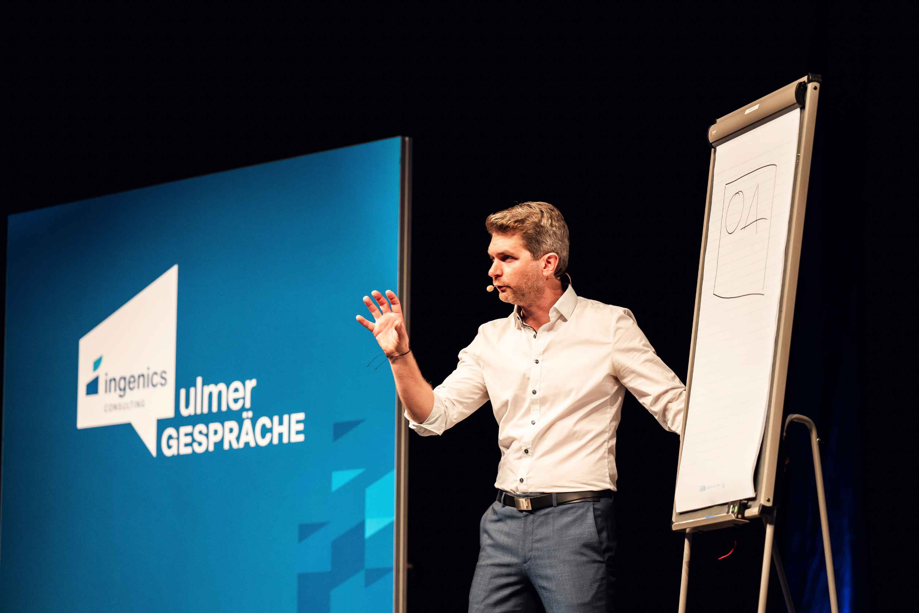 Referierende Person steht neben einem Flipchart und spricht vor Publikum auf einer Veranstaltungsbühne